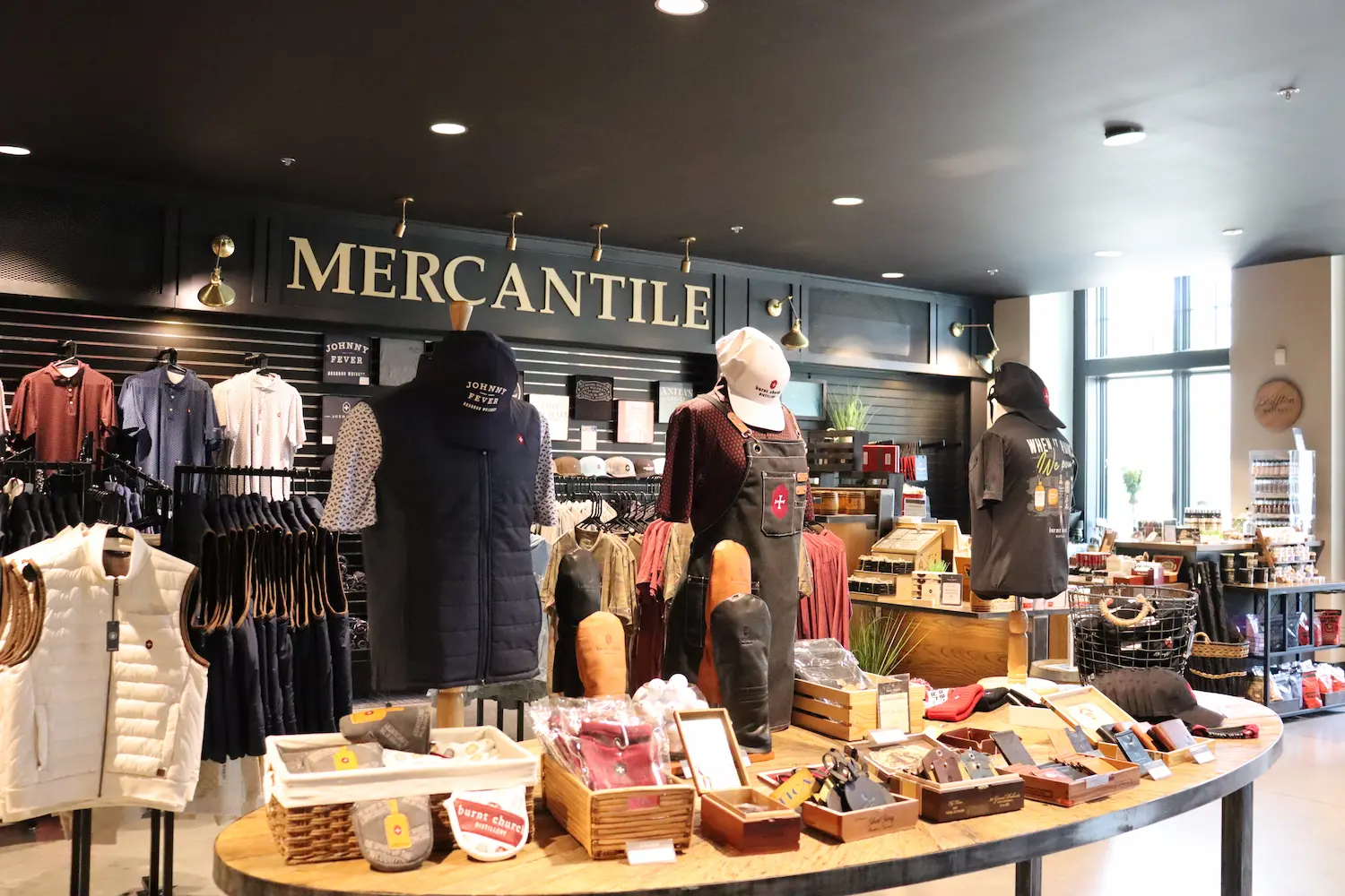 The Mercantile retail shop at Burnt Church Distillery, with branded apparel, hats, and merchandise displayed on racks and a wood table.