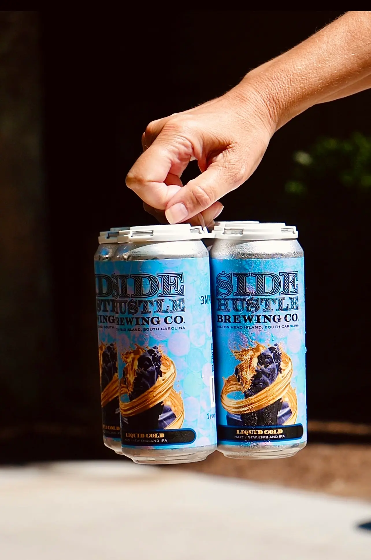 Hand holding a four-pack of Side Hustle Brewing Co. Liquid Gold New England IPA cans.