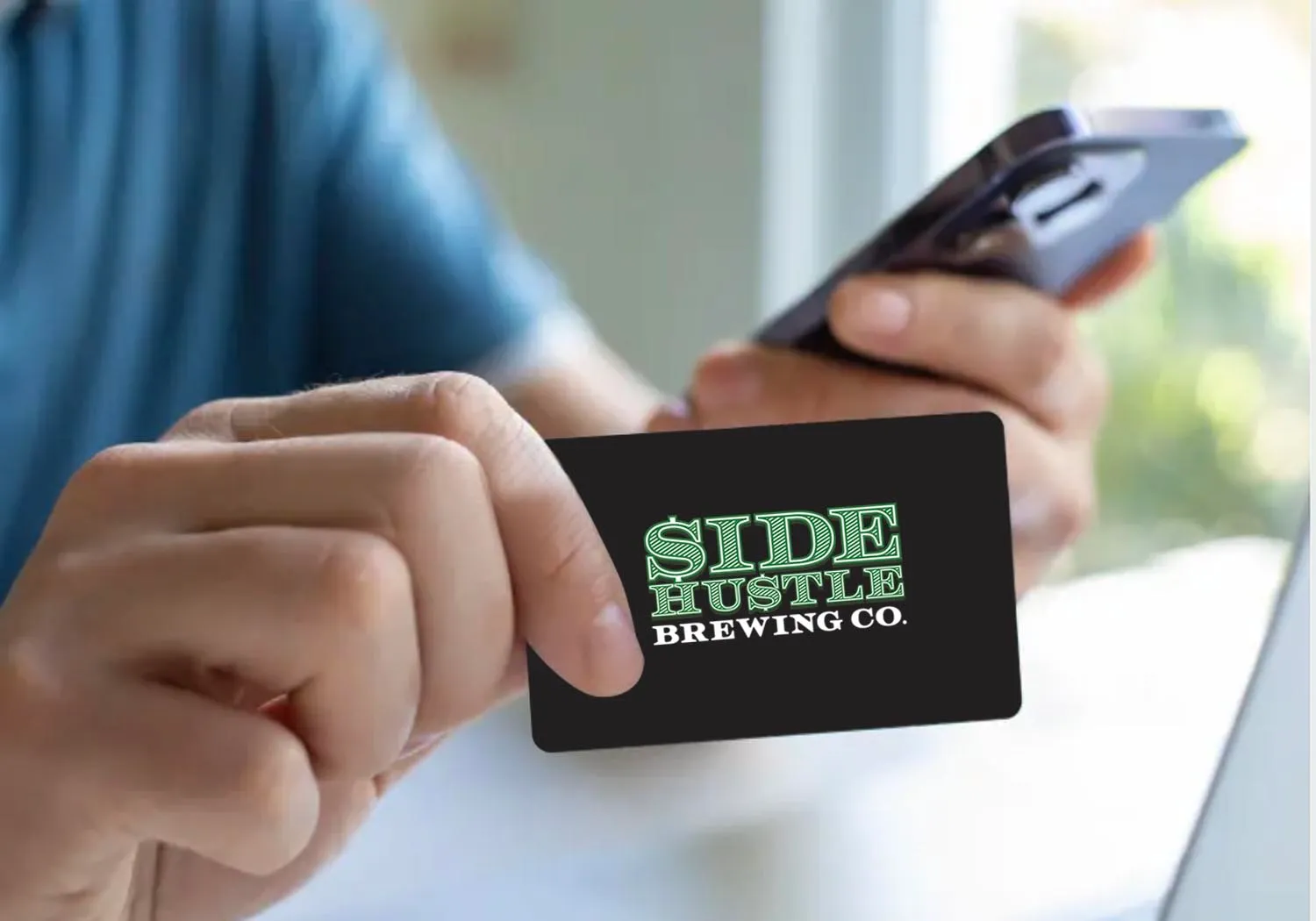A person holding a Side Hustle Brewing Co. gift card in one hand and a phone in the other.
