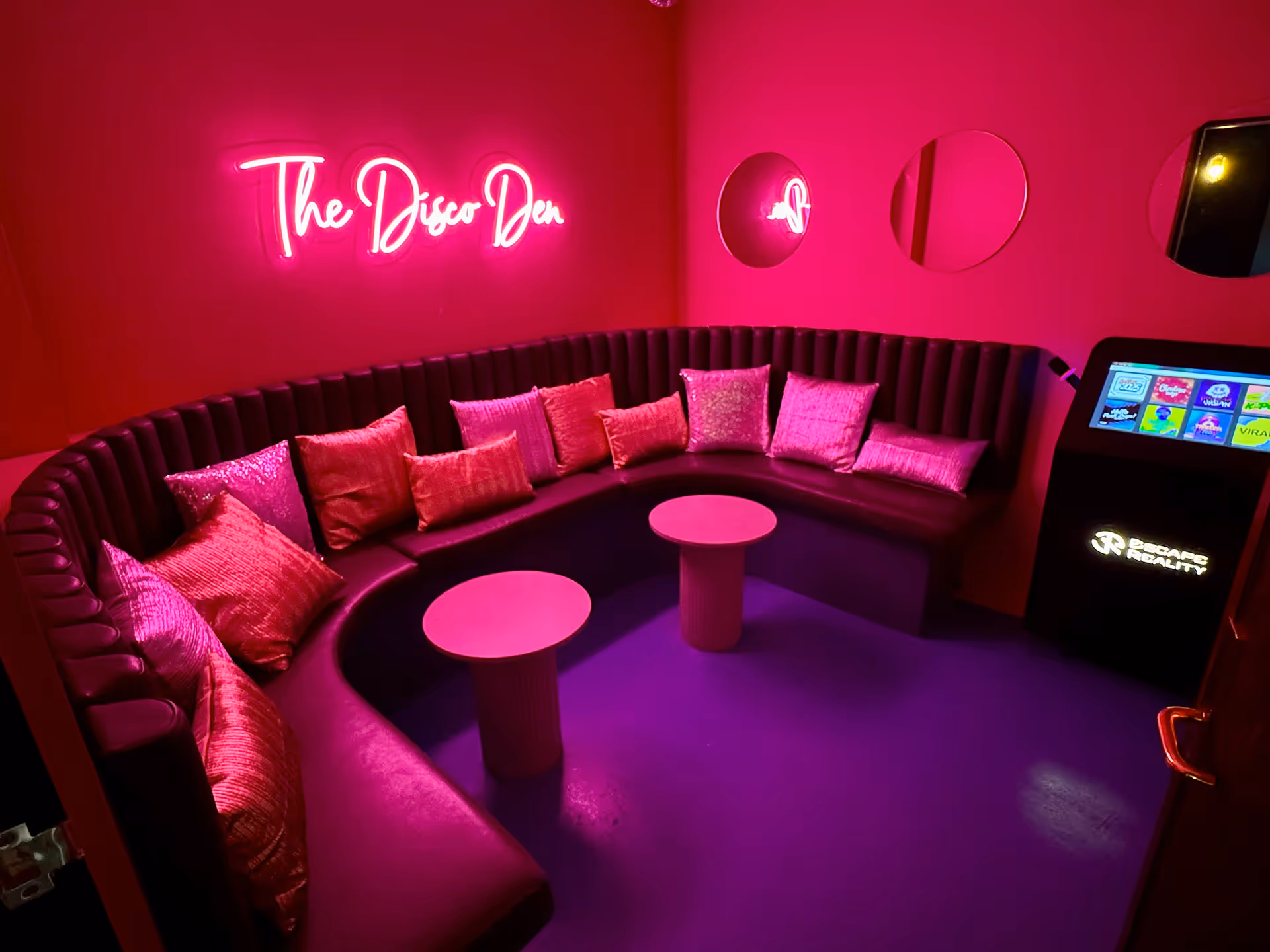 Cozy lounge with curved black seating, pink pillows, two small pink tables, circular mirrors, and neon sign reading 'The Disco Den'.