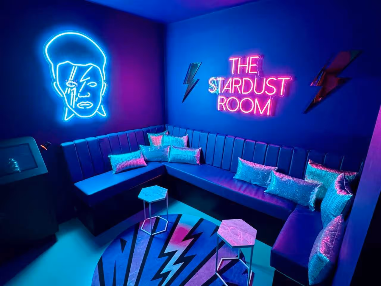 Neon-lit lounge room with a blue and purple color theme, featuring a neon face outline on the wall, a pink neon sign reading 'THE STARDUST ROOM', metallic lightning bolt decorations, cushioned benches, shiny pillows, and hexagonal tables on a patterned rug.