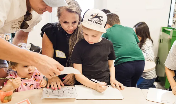Adults helping young children with writing and drawing activities at a classroom table.