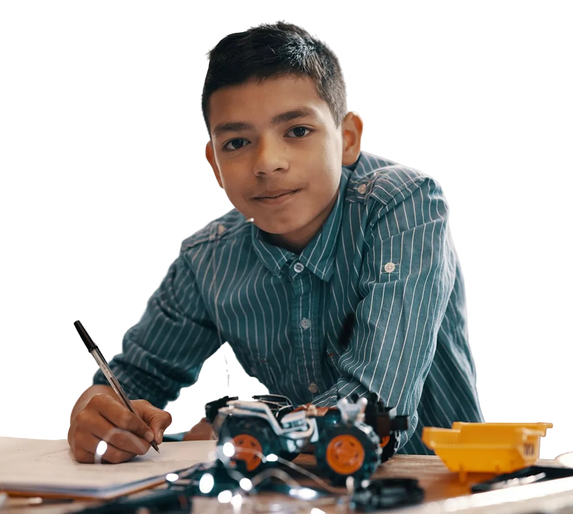Boy wearing a striped shirt writing on paper with toy vehicles in front of him on a table.