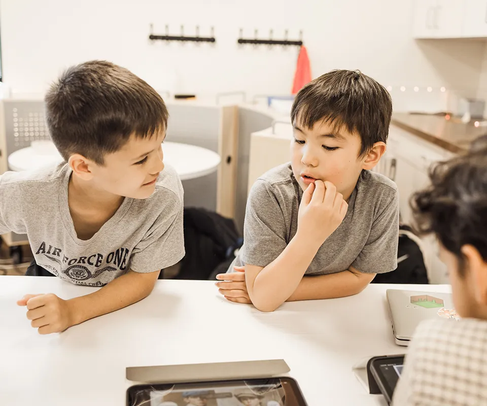 Two boys leaning on a table watching a tablet screen with concentration in a modern classroom.