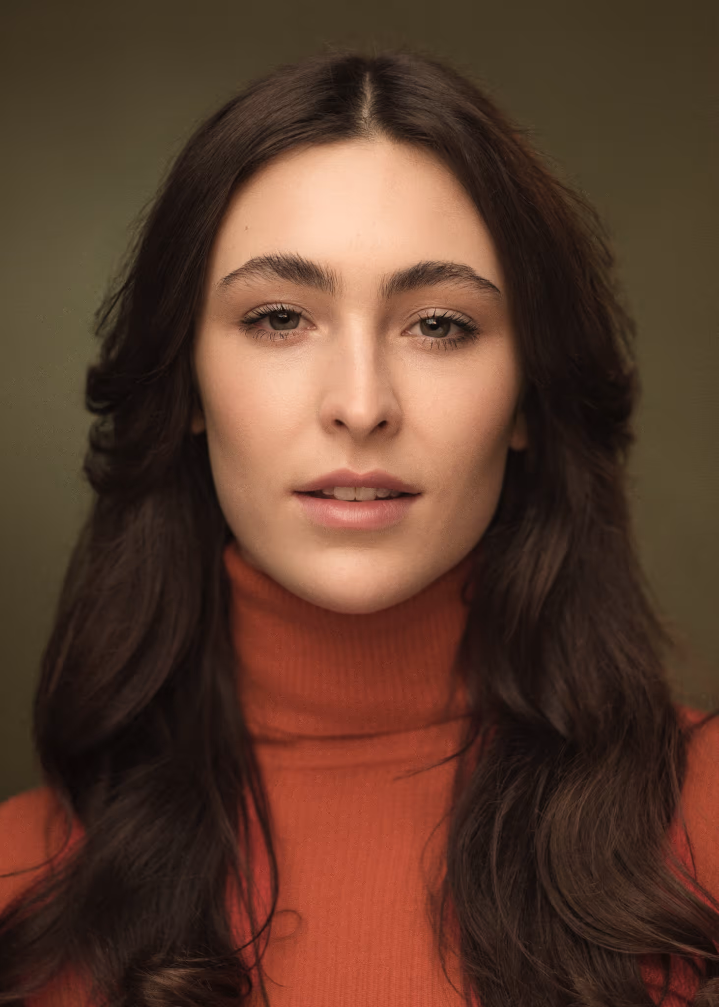 Close-up portrait of Yaman Kelemet with long dark hair wearing a burnt orange turtleneck sweater against an olive green background.