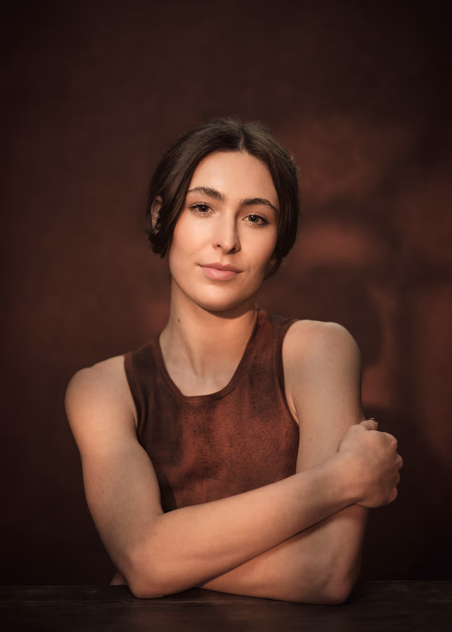 Yaman kelemet with dark hair and a sleeveless brown top sitting with arms crossed against a dark brown background.