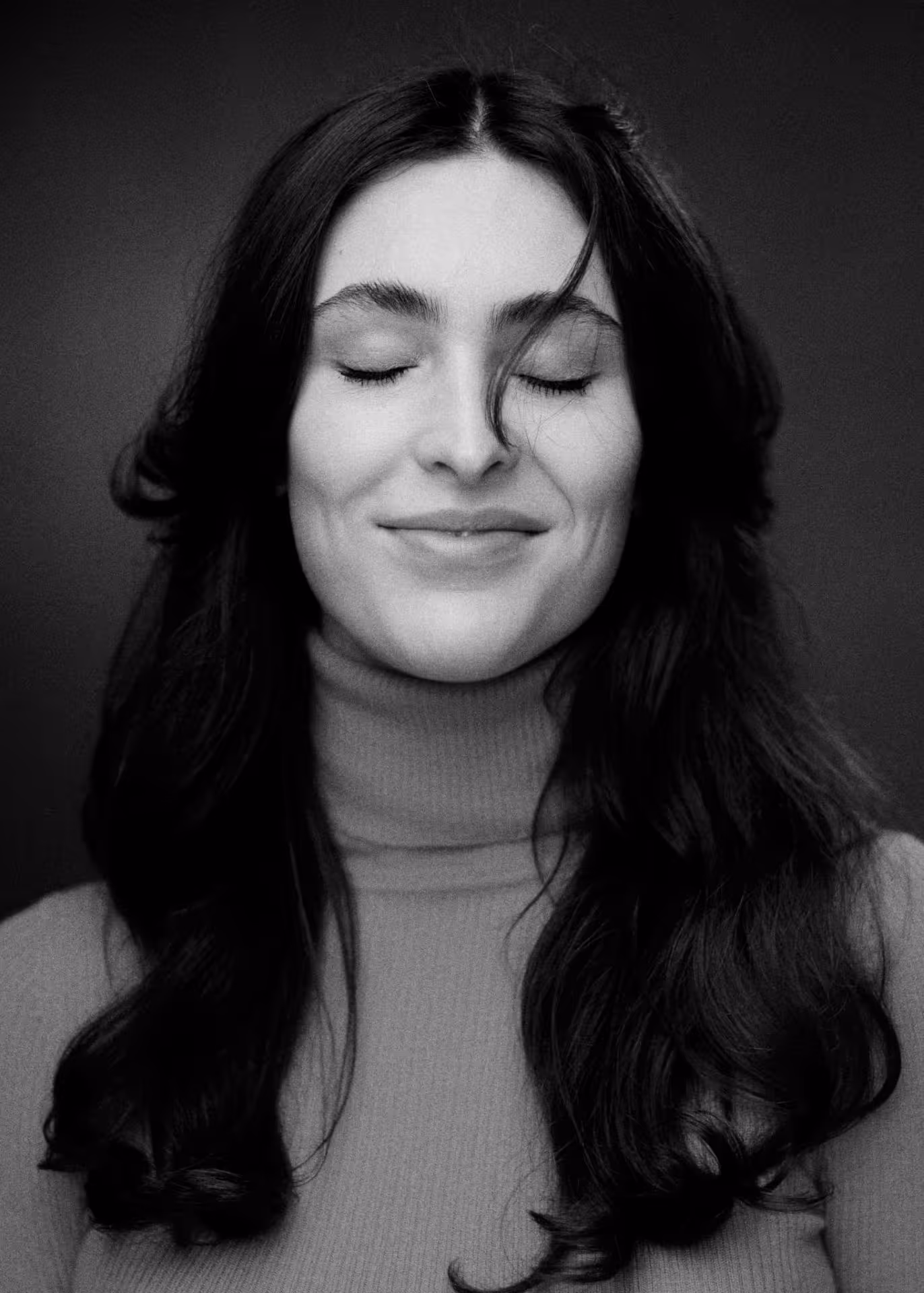Black and white portrait of Yaman Kelemet with long dark hair, eyes closed and a gentle smile, wearing a turtleneck sweater.