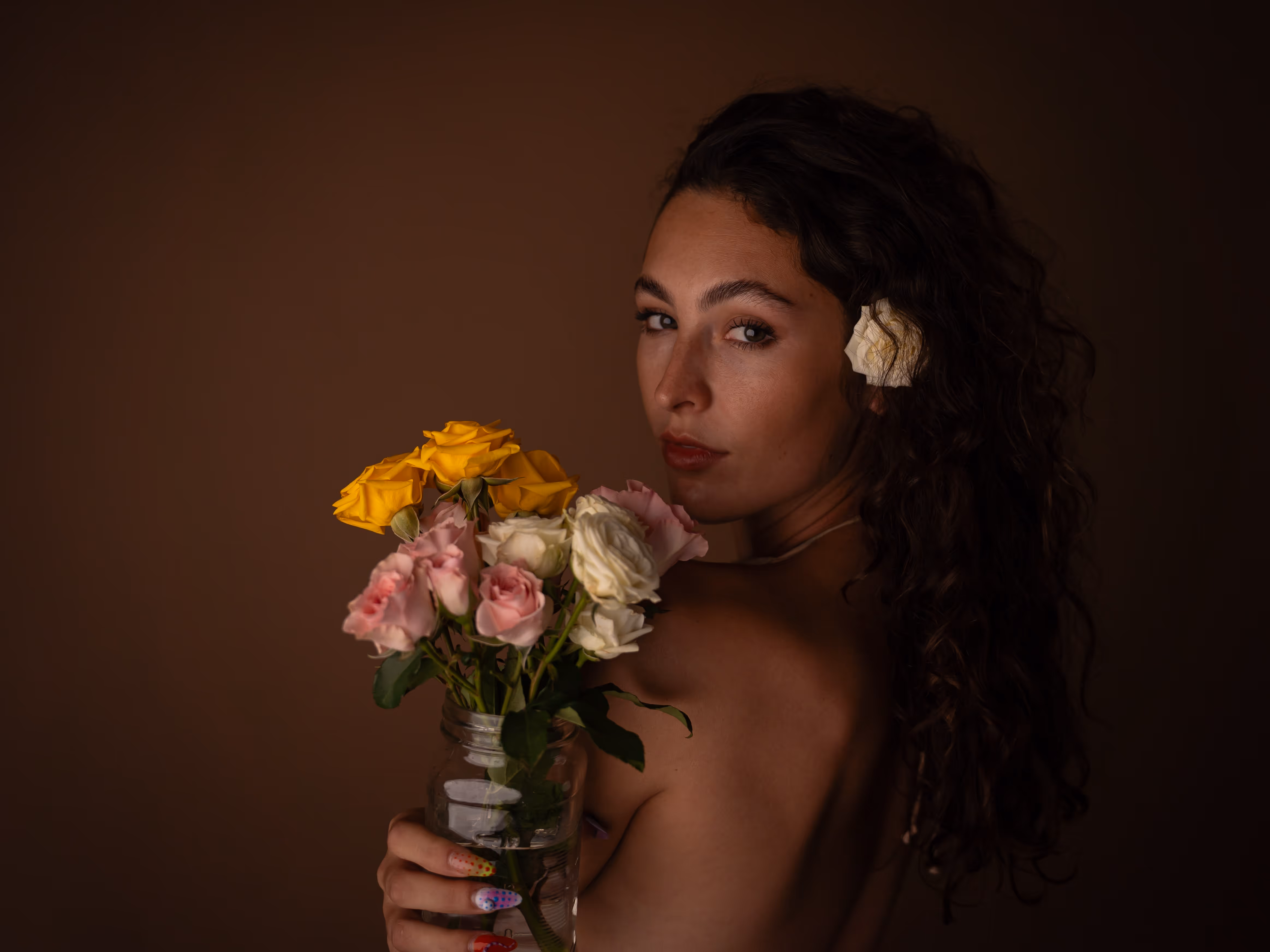 Yaman Kelemet with curly dark hair and flower tucked behind ear holding a bouquet of yellow, pink, and white roses in a glass jar against a brown background.
