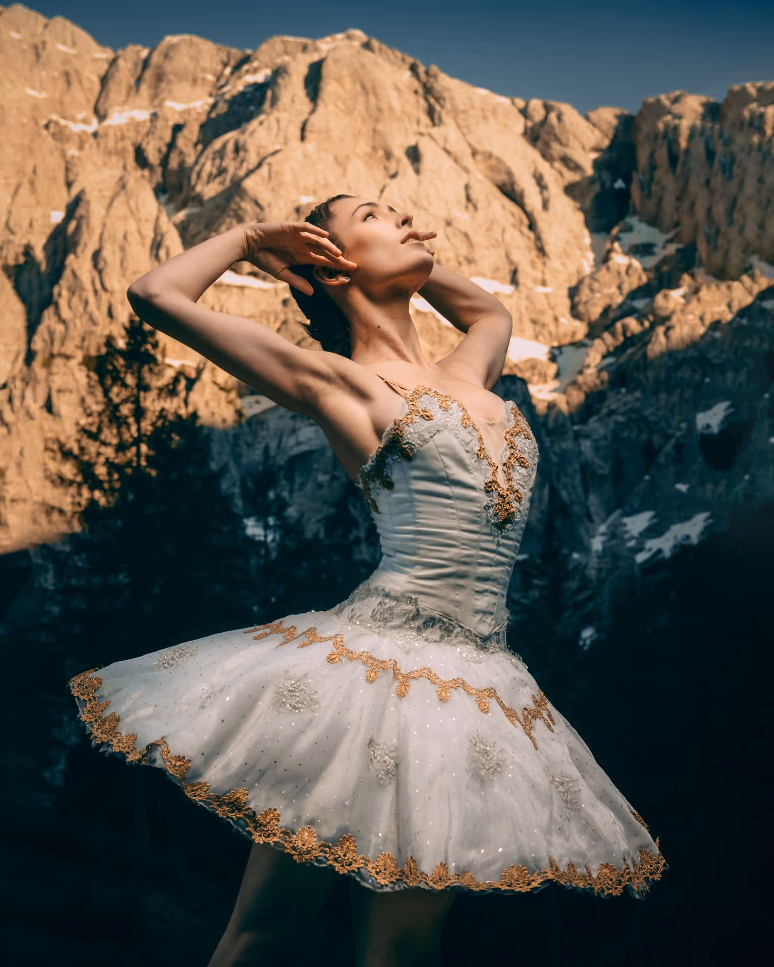 Yaman Kelemet in a white tutu with gold embroidery posing gracefully against a rocky mountain backdrop.