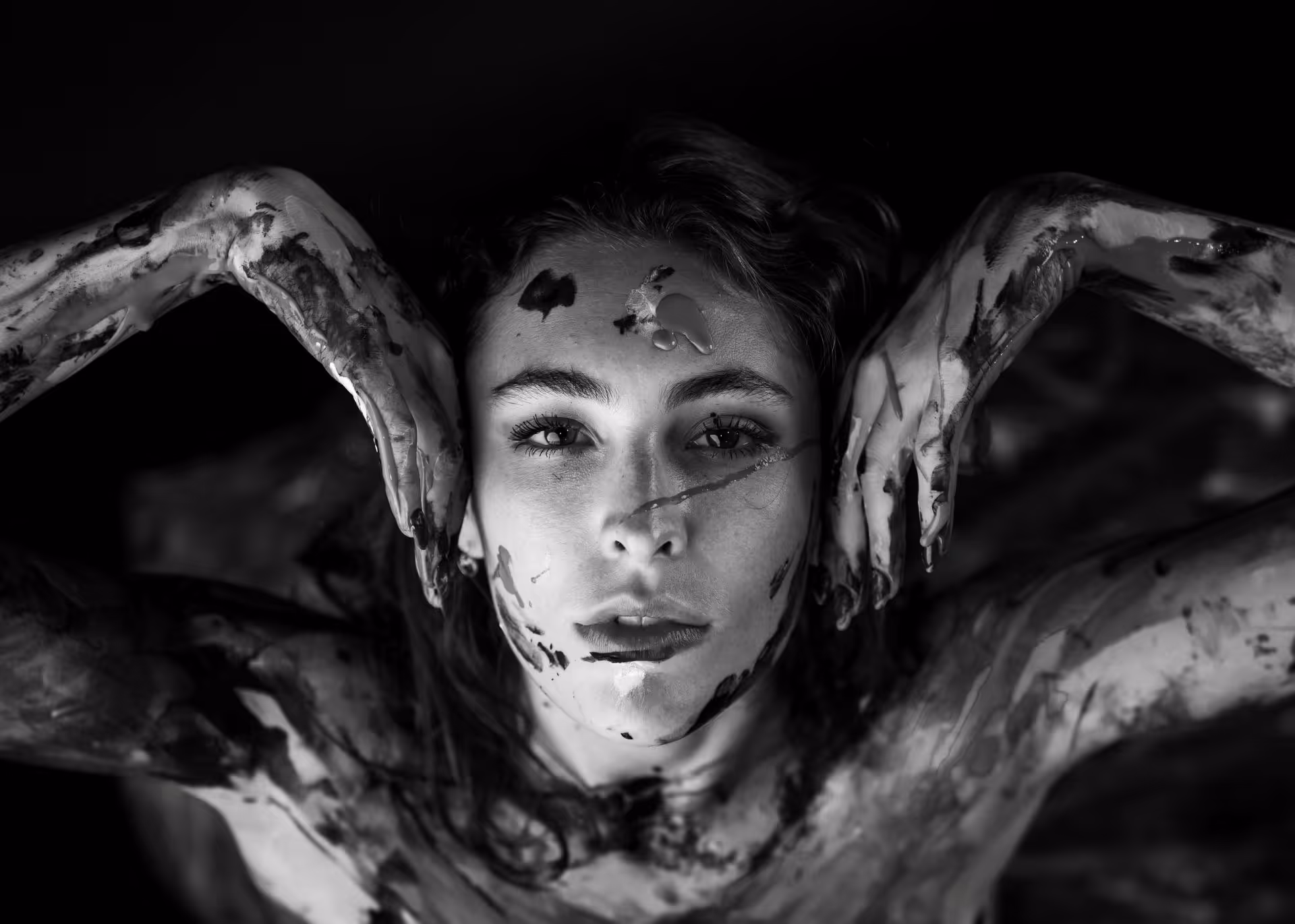 Black and white portrait of Yaman Kelemet with paint smeared on her face and arms, posing with hands framing her face.
