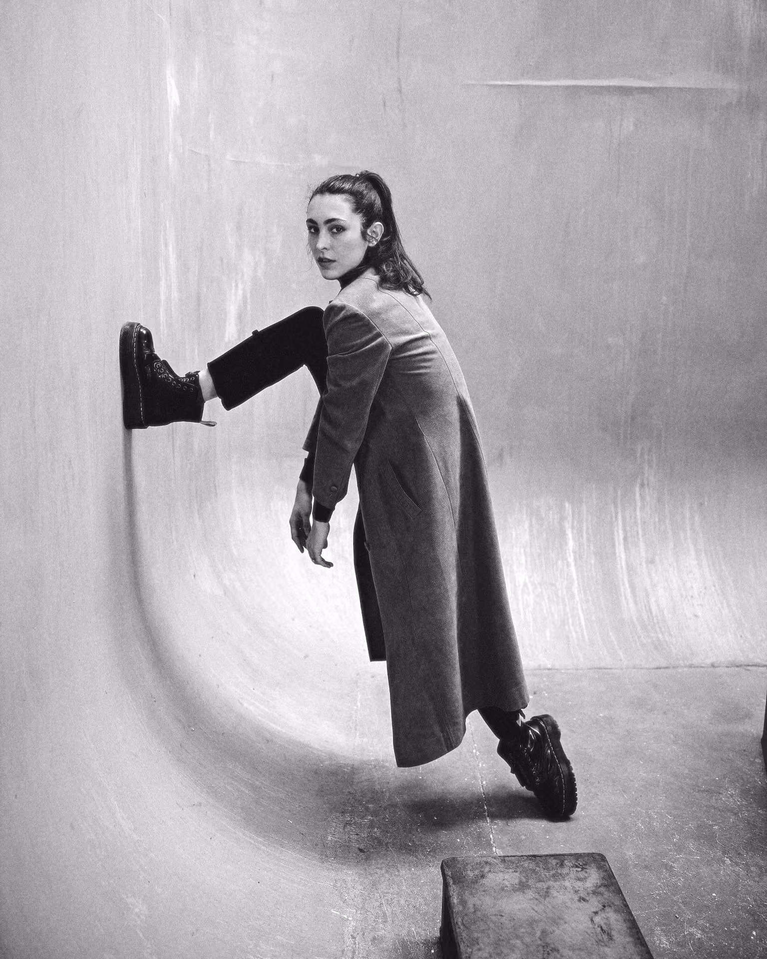 Black and white photo of Yaman Kelemet in a long coat posing with one leg against a curved wall.