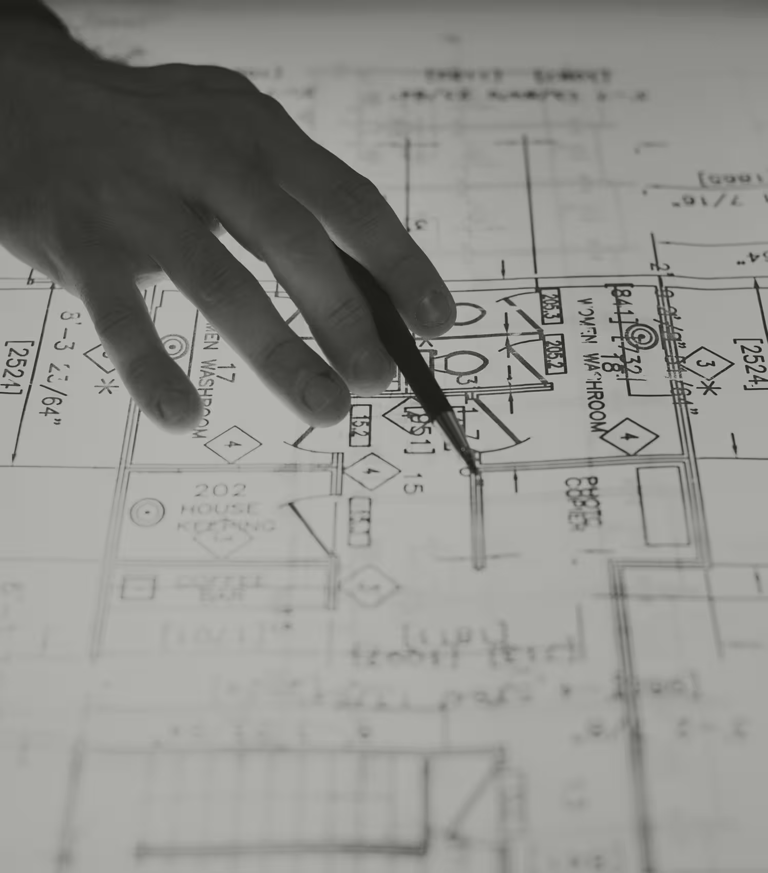 Hand holding a pen pointing to a section of an architectural blueprint showing room labels and measurements.