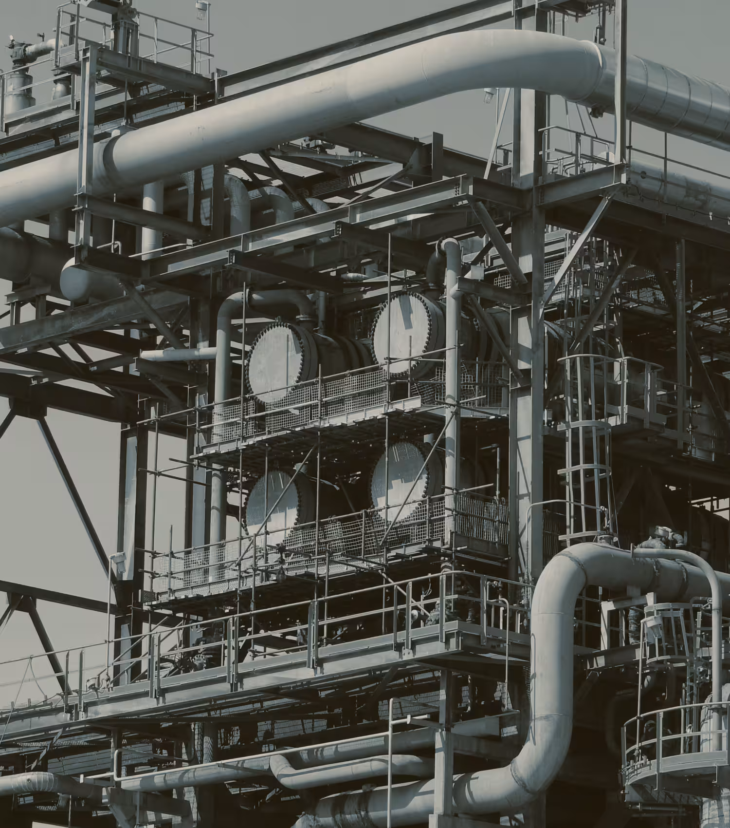 Close-up of industrial facility with large metal pipes, cylindrical tanks, and scaffolding structures.