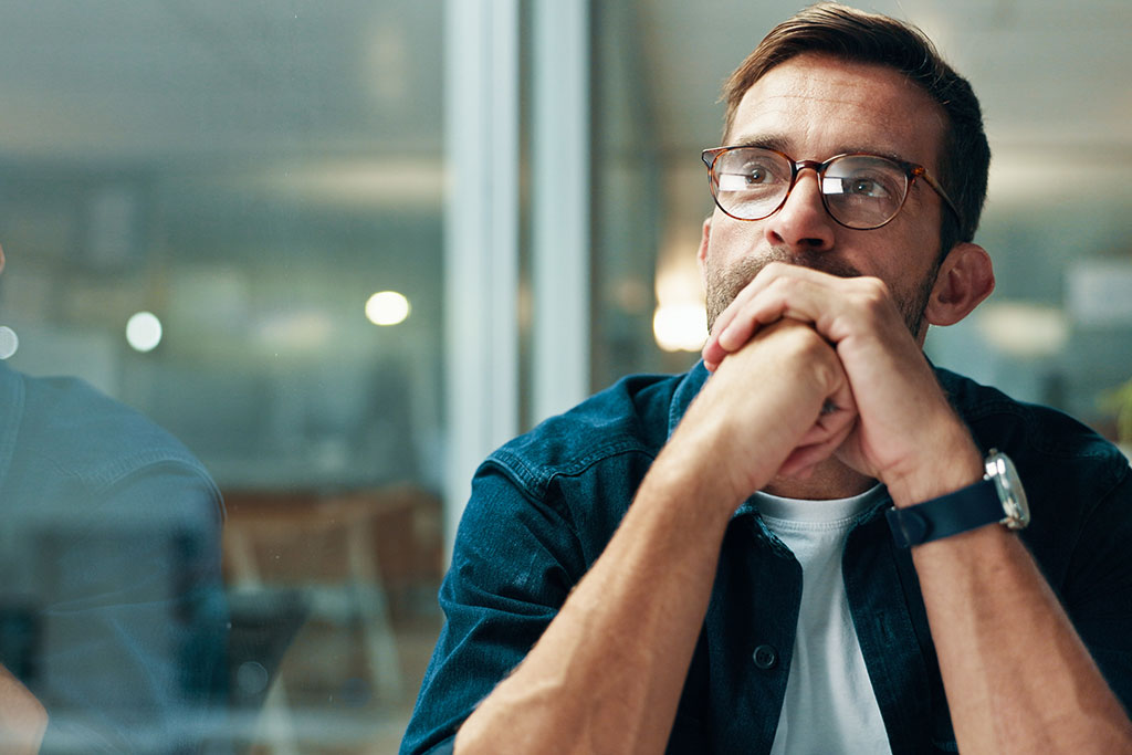A thoughtful man wearing glasses sits with his hands clasped under his chin, looking off-camera.