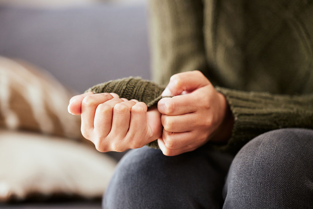 A close-up of a person's hands tightly clenching and pulling at the sleeve of an olive green knit jumper, suggesting anxiety or tension.