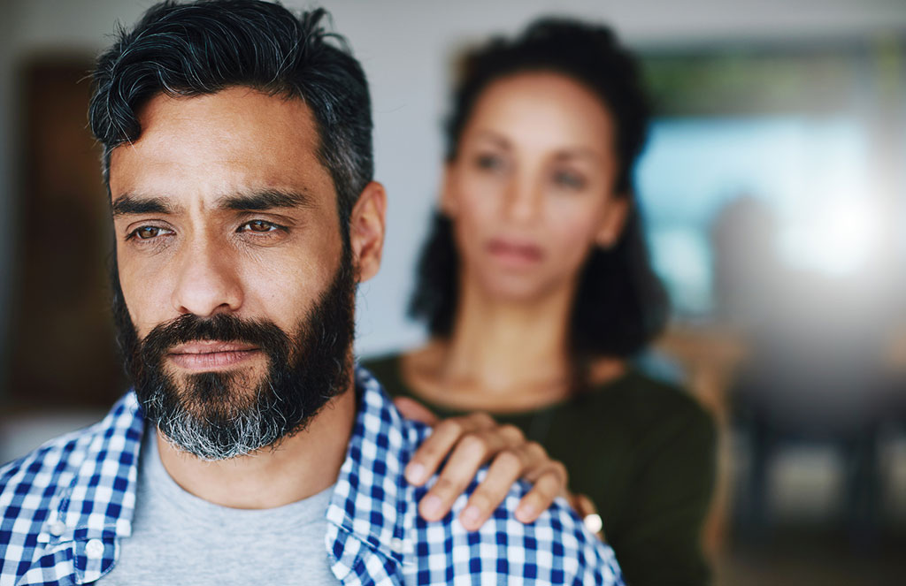 A close-up of a distressed man with a beard looking away while a woman places a supportive hand on his shoulder in the background.
