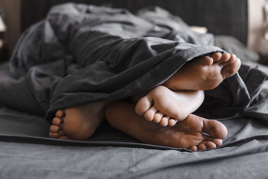 Two pairs of feet intertwined and poking out from under a dark grey duvet on a bed.
