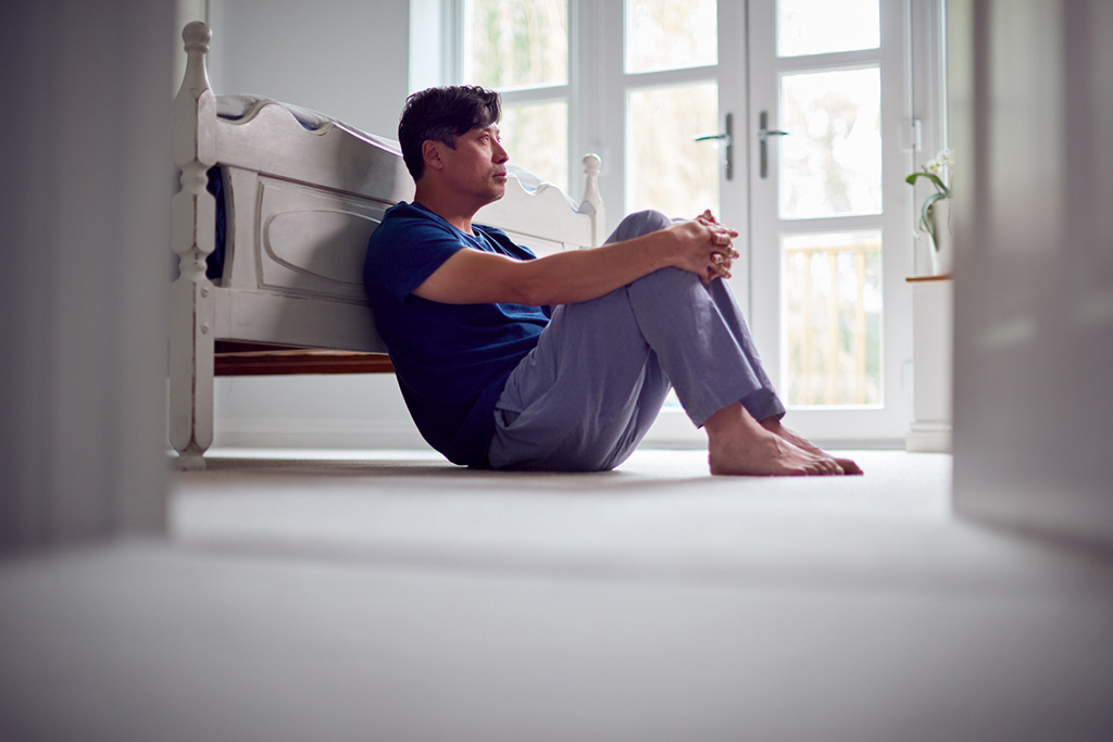 A man in pajamas sits on the bedroom floor leaning against a bed, looking thoughtfully toward a window.