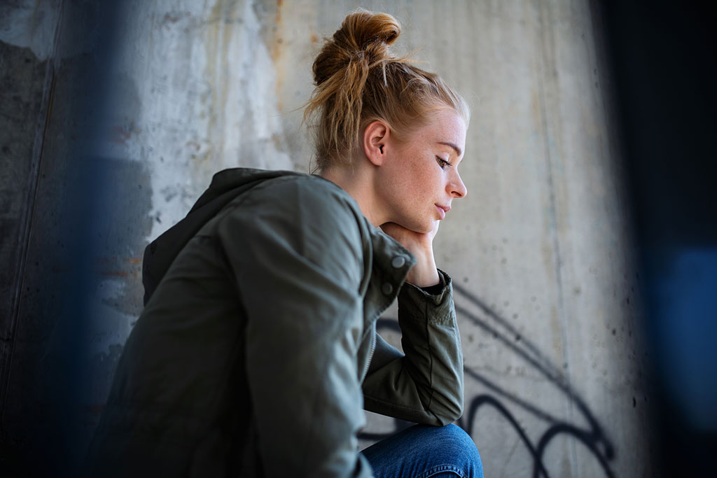 A young woman with red hair in a bun sits in profile against a concrete wall, looking down thoughtfully.