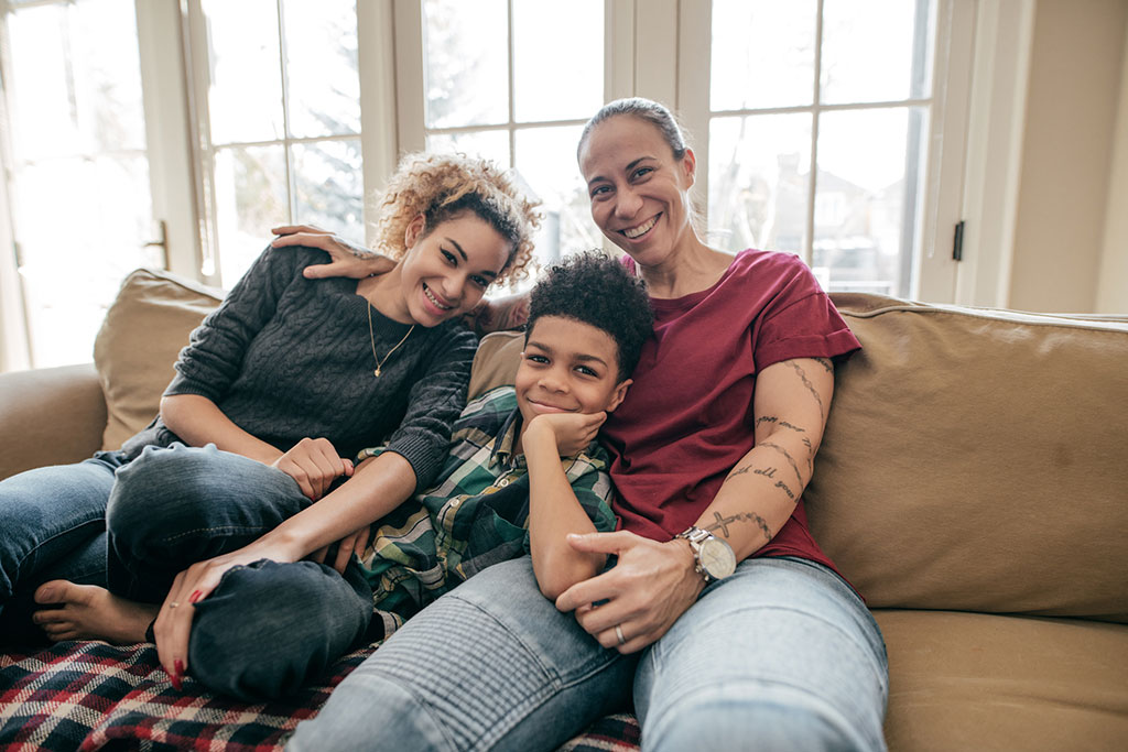 A smiling family of three—two women and a young boy—sitting closely together on a living room sofa.