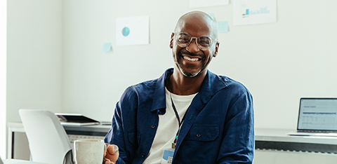 A smiling, bald Black man wearing glasses and a blue button-down shirt sits at an office desk, holding a coffee mug.