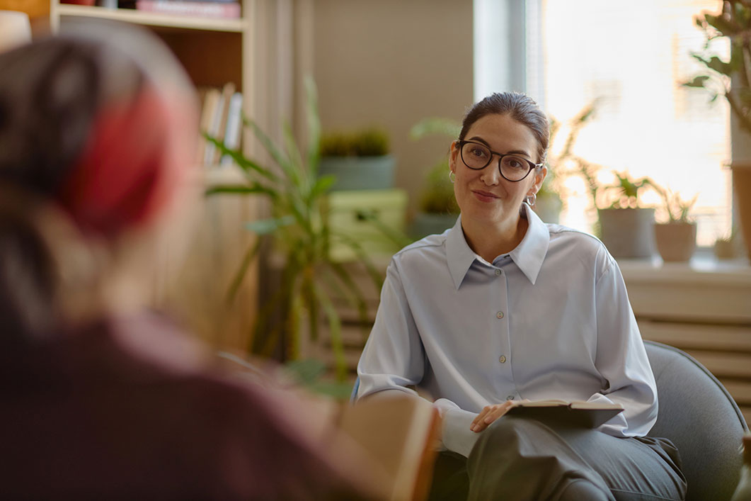 A female therapist wearing glasses and a light blue button-down shirt sits in a chair, listening intently to a client whose back is partially visible in the foreground.