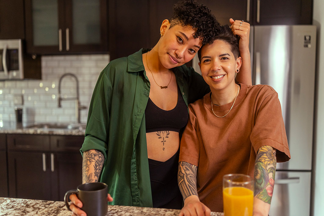 A smiling couple with tattoos stands close together behind a kitchen counter, holding a coffee mug and a glass of orange juice.