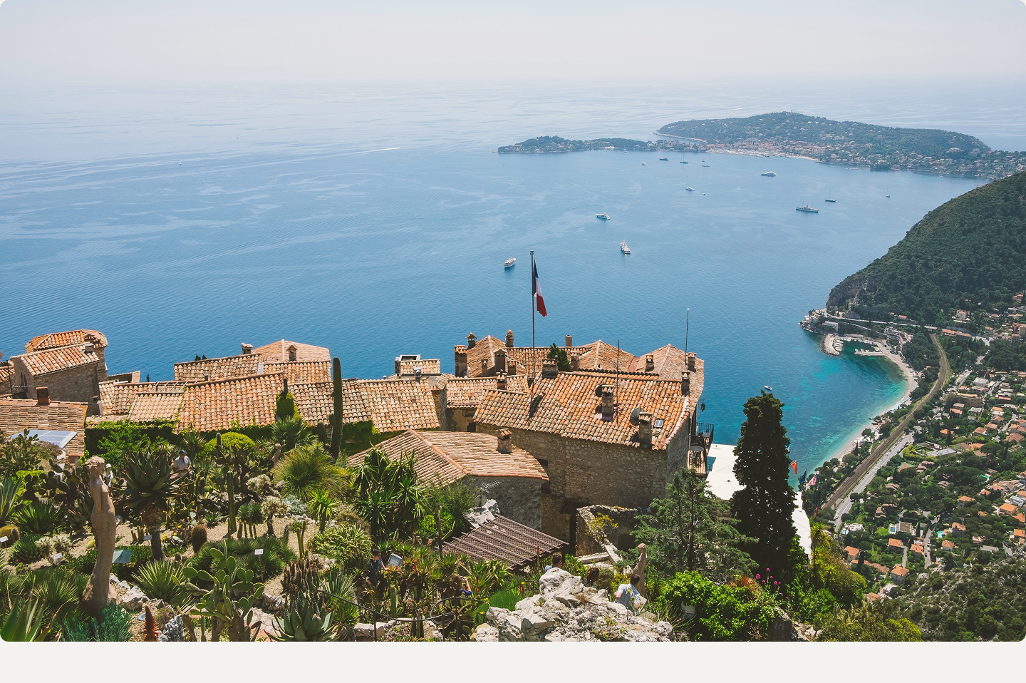 View of terracotta rooftops, greenery, and a French flag overlooking a coastal bay with boats and island in the distance.