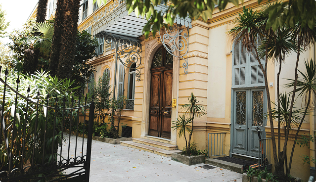 Elegant yellow building entrance with a wooden door, ornate ironwork, and surrounding tropical plants.