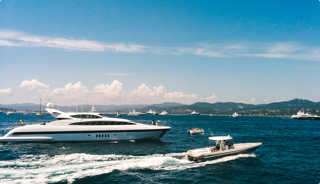 White luxury yachts and a speedboat cruising on blue ocean water with mountainous coastline under a clear sky.