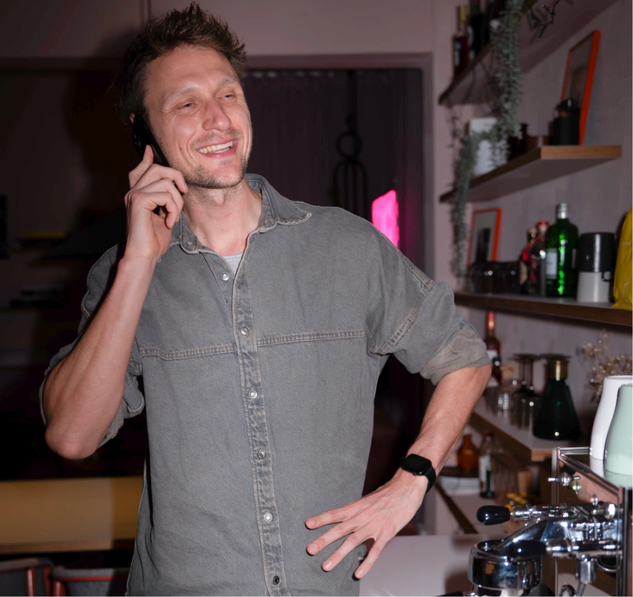 Mann mit grauem Hemd telefoniert lächelnd in einer Bar mit Kaffeemaschine und Regalen im Hintergrund.