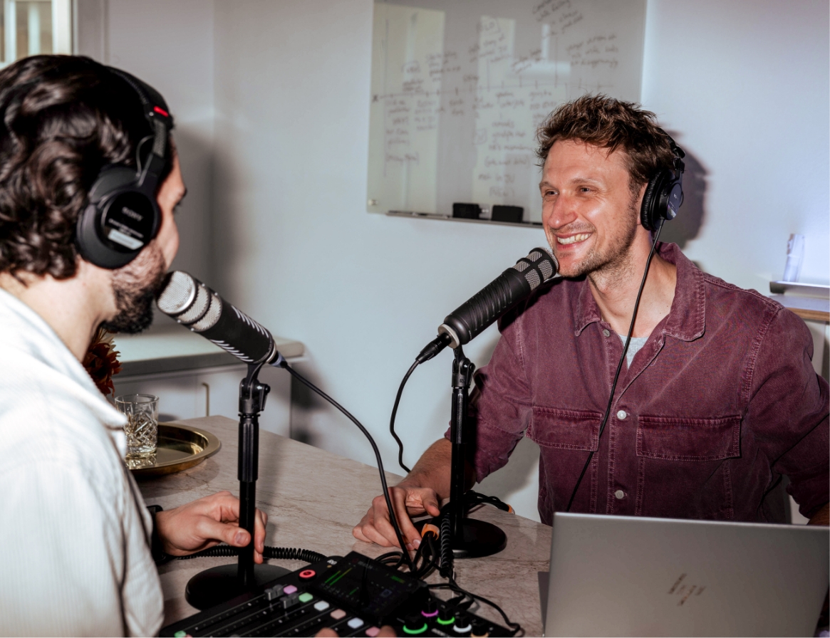 Zwei Männer im Gespräch, tragen Headsets und sprechen in Mikrofone, mit Podcast-Equipment vor sich.
