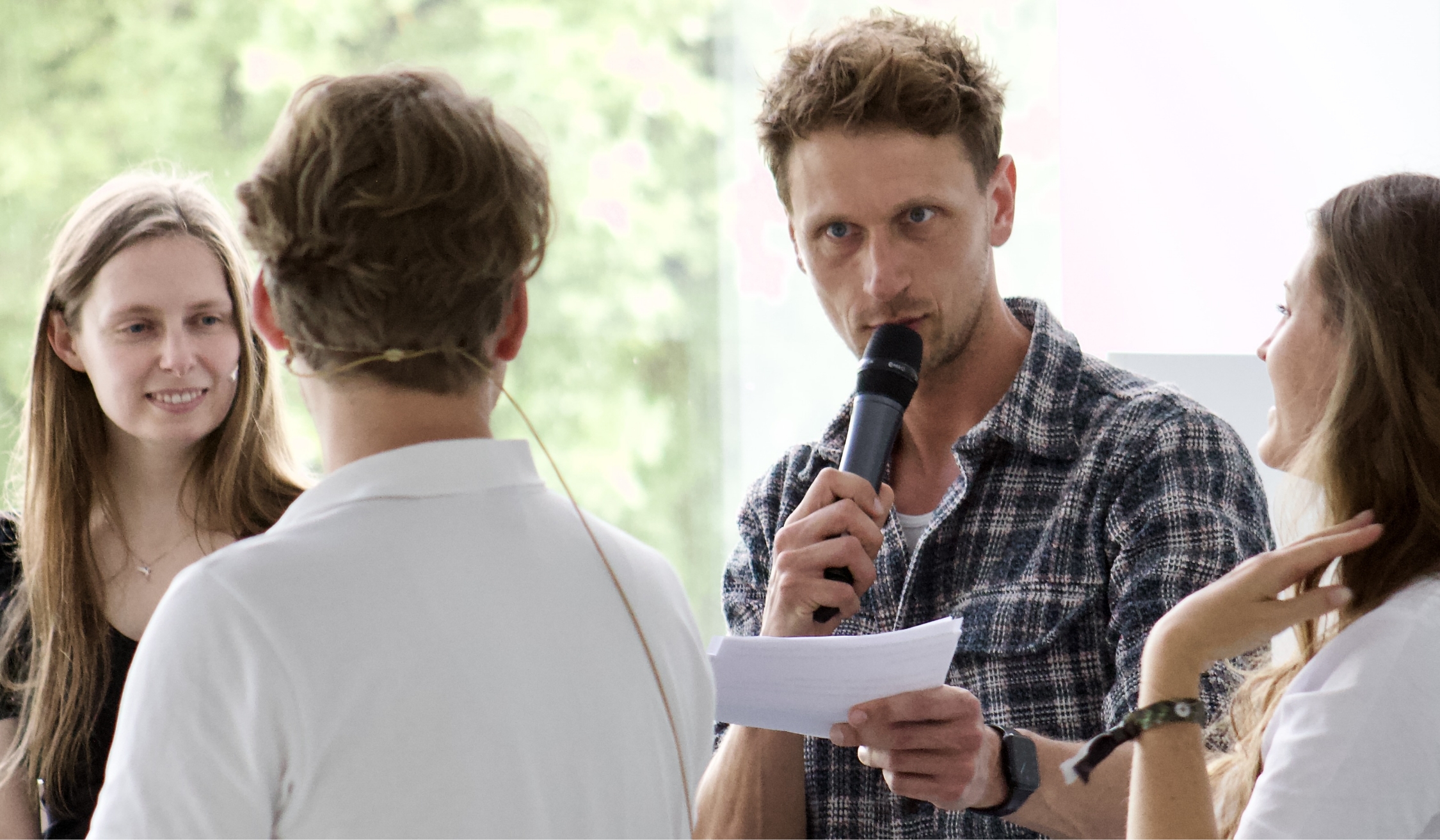 Mann spricht in Mikrofon, drei Personen hören ihm in einer hellen Umgebung zu.