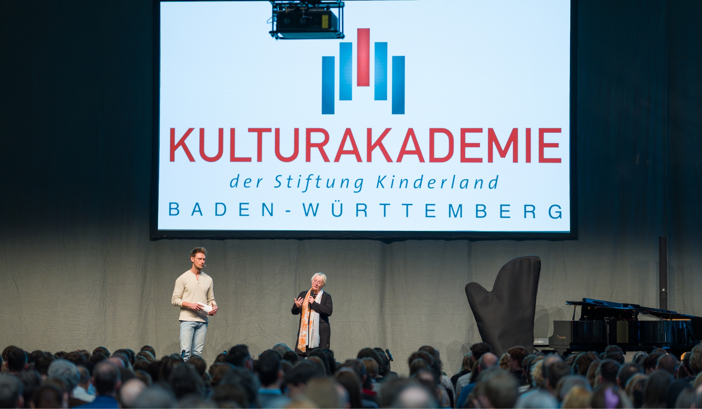 Zwei Redner auf Bühne vor Publikum mit großem Bildschirm der Kulturakademie Baden-Württemberg.
