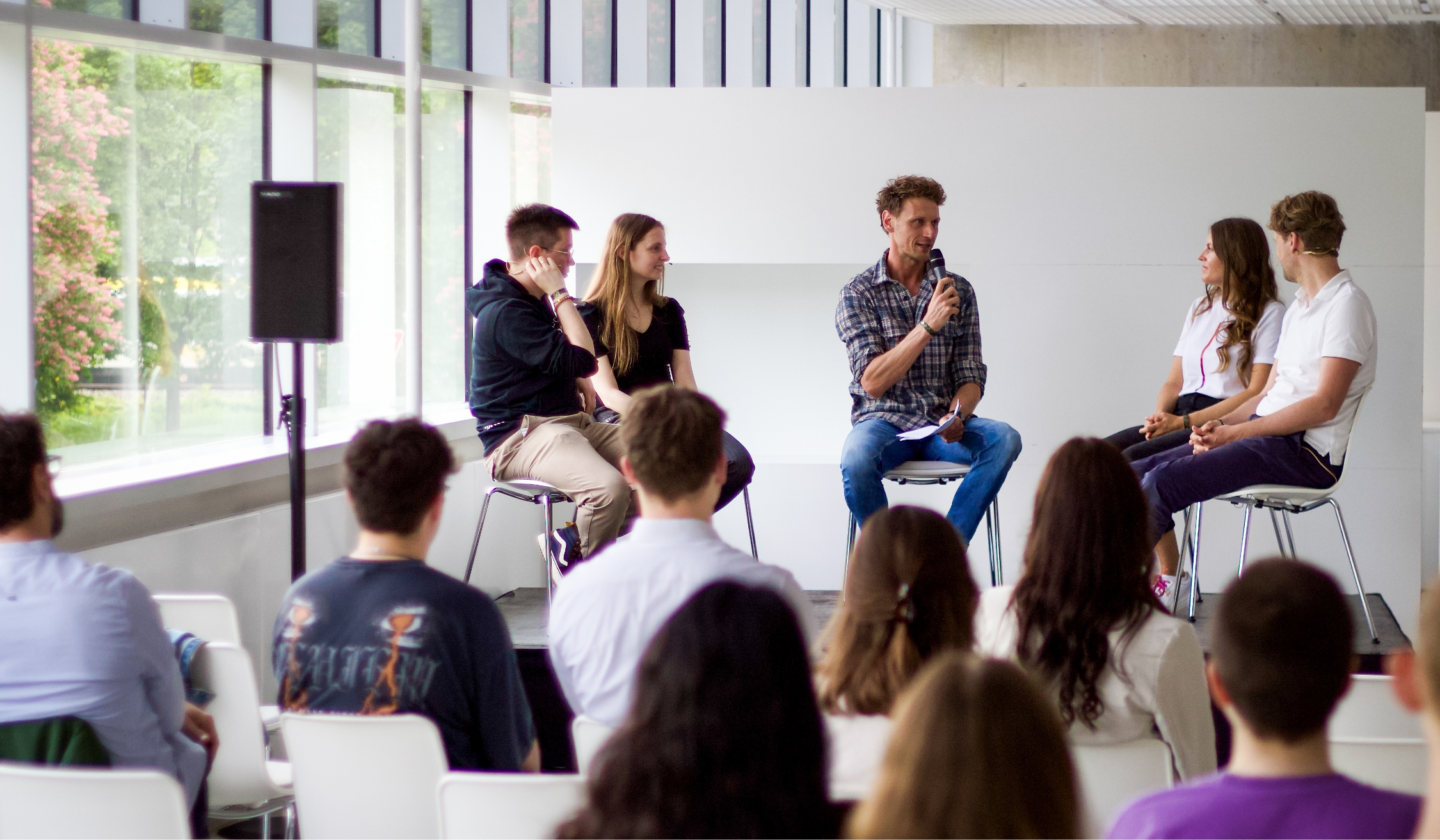 Podiumsdiskussion mit fünf Personen, eine Person spricht in ein Mikrofon vor sitzendem Publikum.