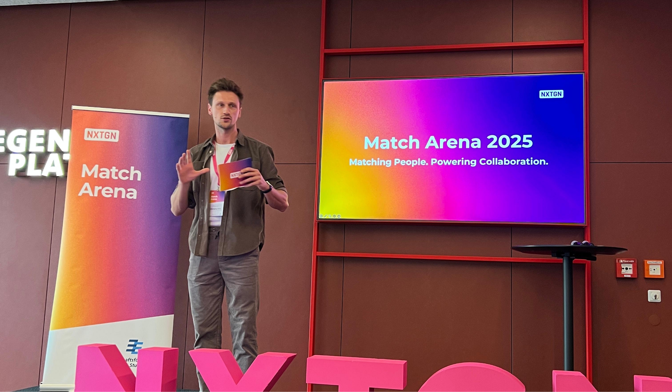 Mann hält Präsentation vor Match Arena Banner und Bildschirm mit Text Match Arena 2025.