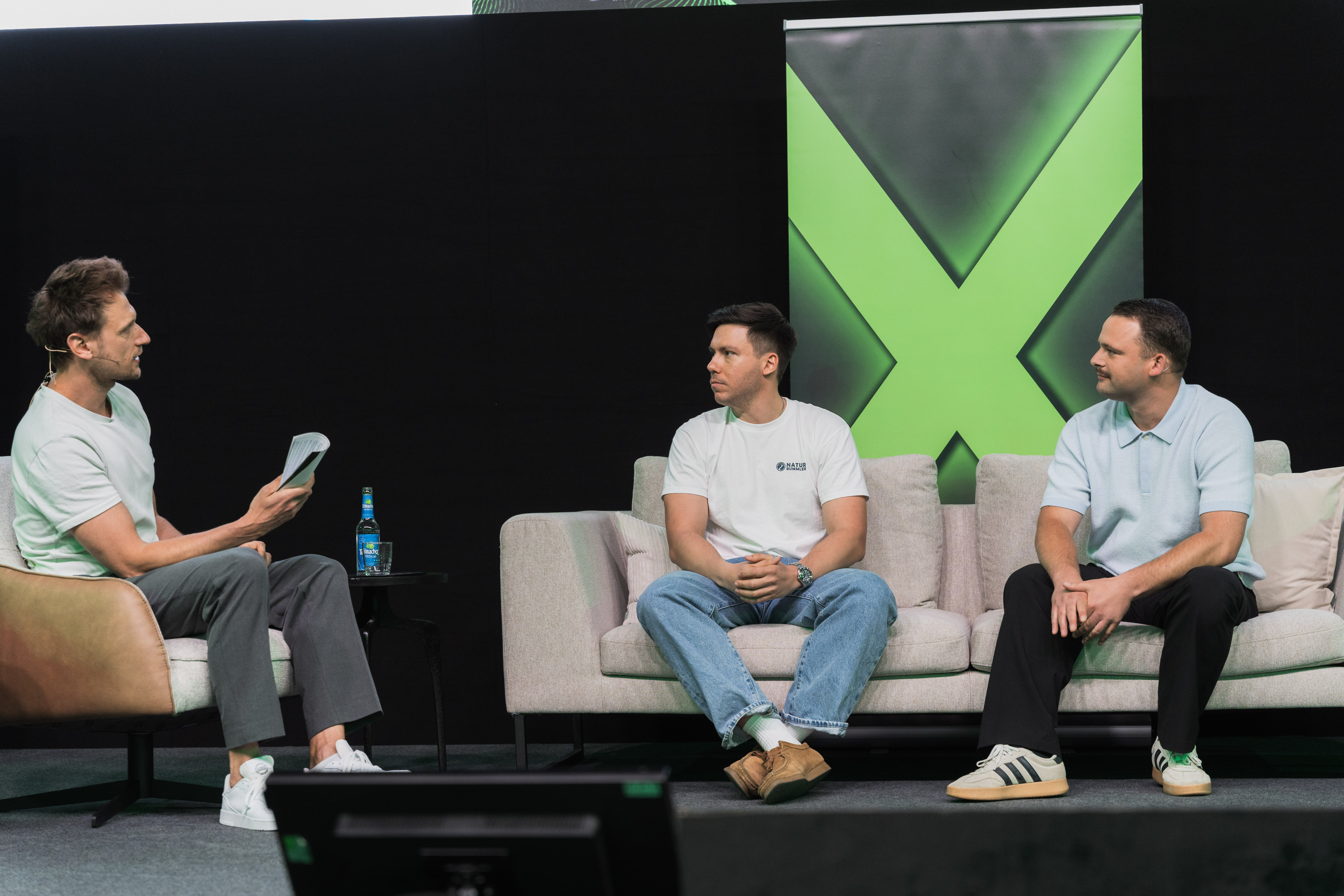 Drei Männer führen eine Podiumsdiskussion auf der Bühne mit grünem X-Banner im Hintergrund.