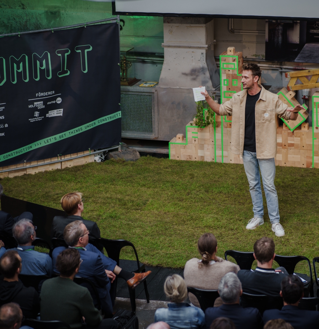 A man speaks on a grassy stage to an audience seated in black chairs during an outdoor event.