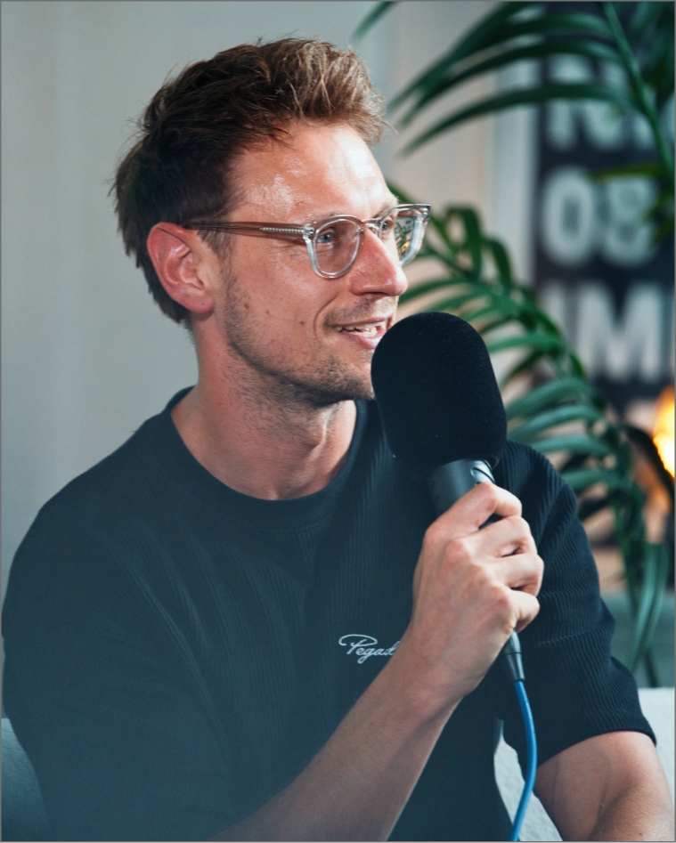 Mann mit Brille hält Mikrofon und spricht, grüner Zimmerpflanze im Hintergrund.