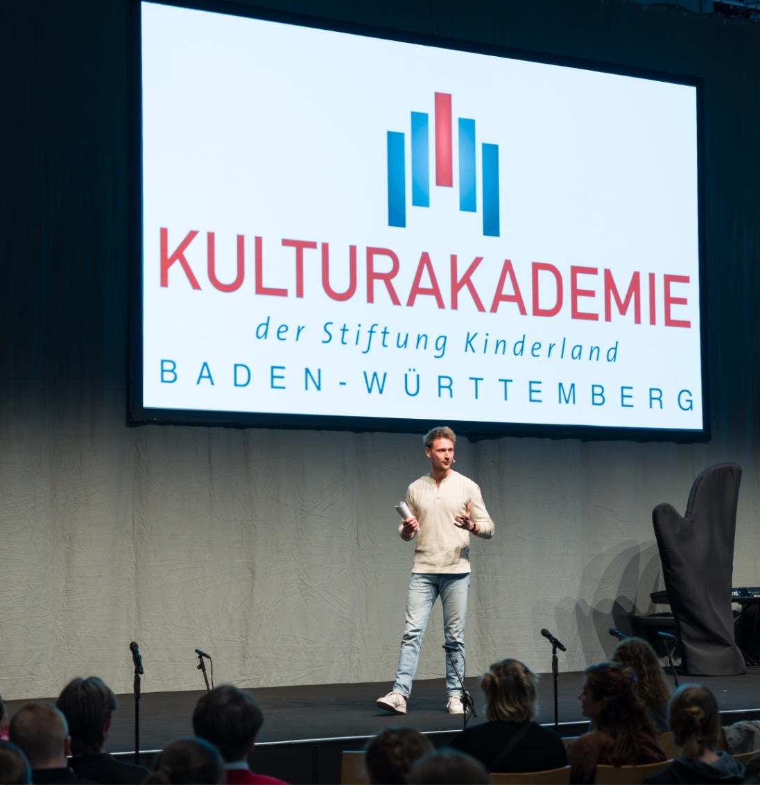 Mann spricht auf Bühnenpodium vor Publikum, auf großem Bildschirm steht Kulturakademie Baden-Württemberg.