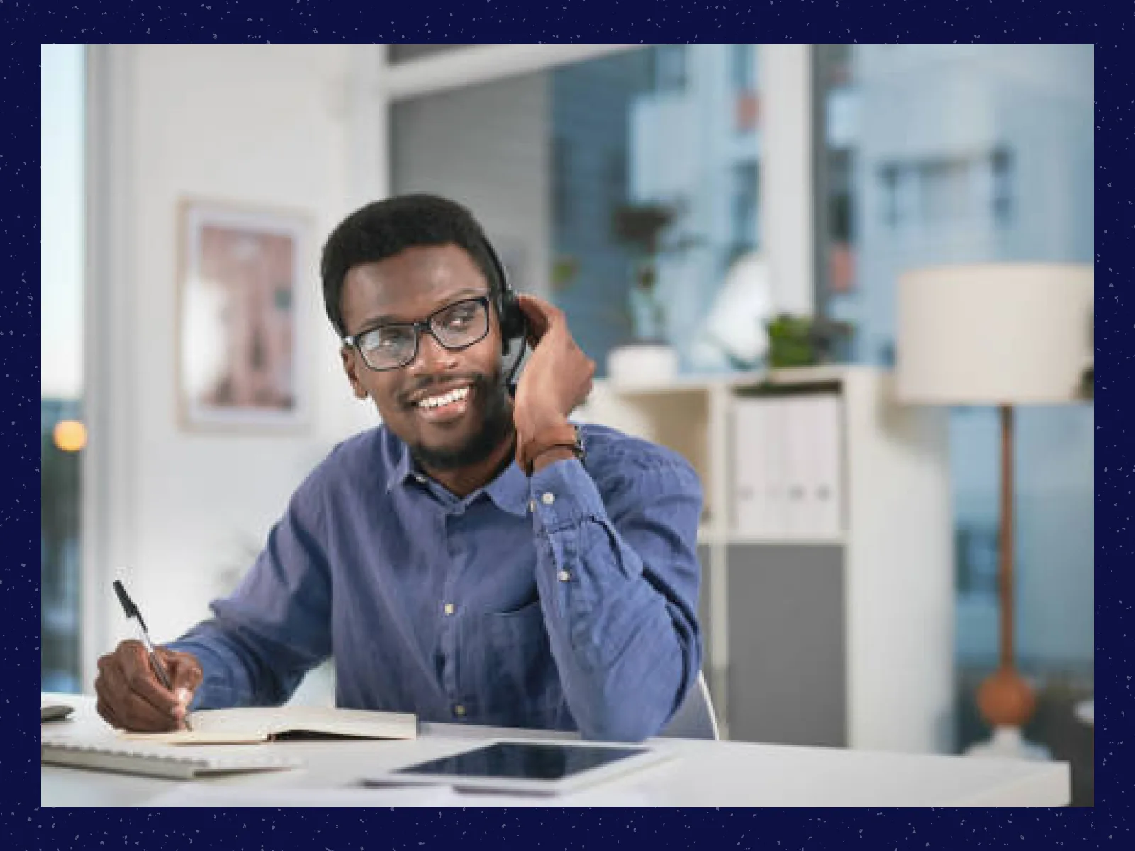 Smiling man wearing glasses and headset writing in notebook at desk with tablet and keyboard.