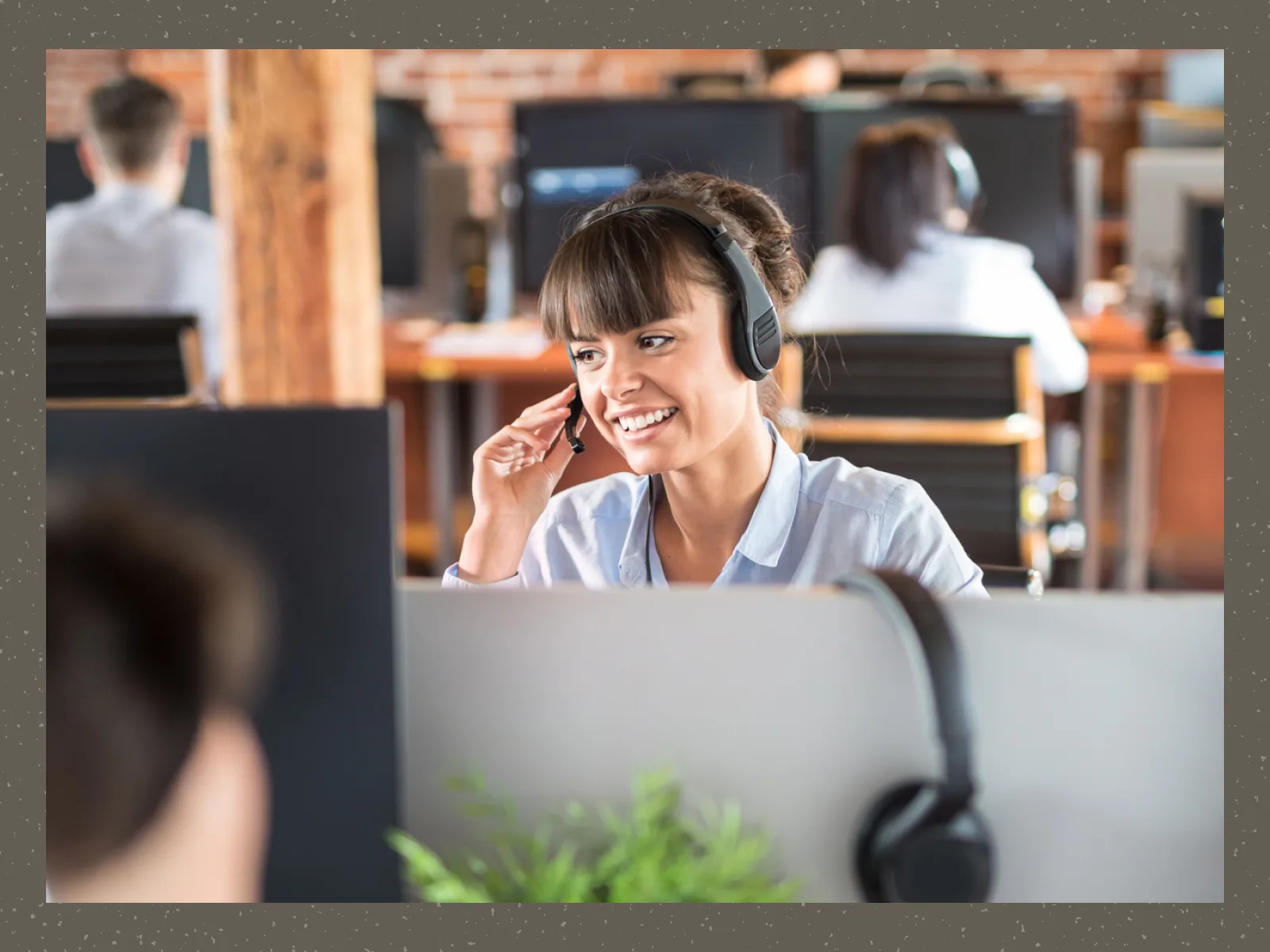 Smiling woman wearing headset talking at her desk in a busy open office environment.