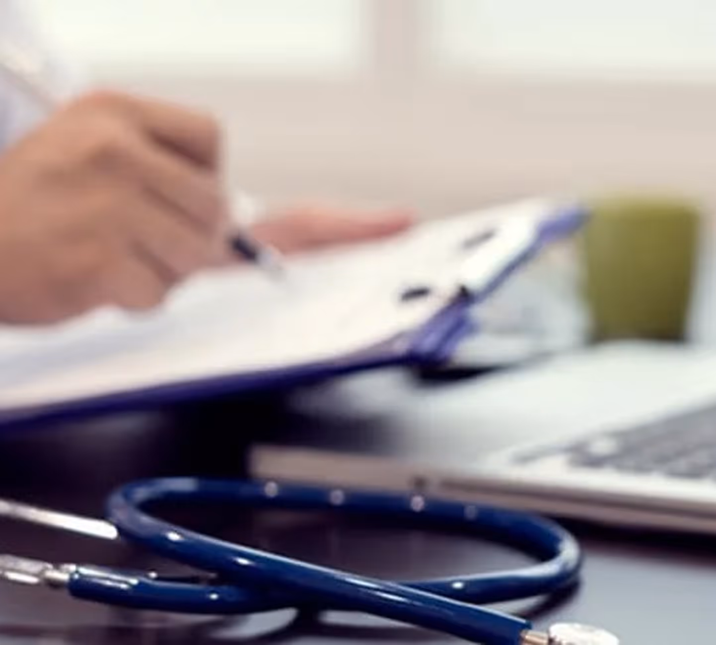 Blue stethoscope on desk with blurred doctor writing on clipboard and laptop in background.