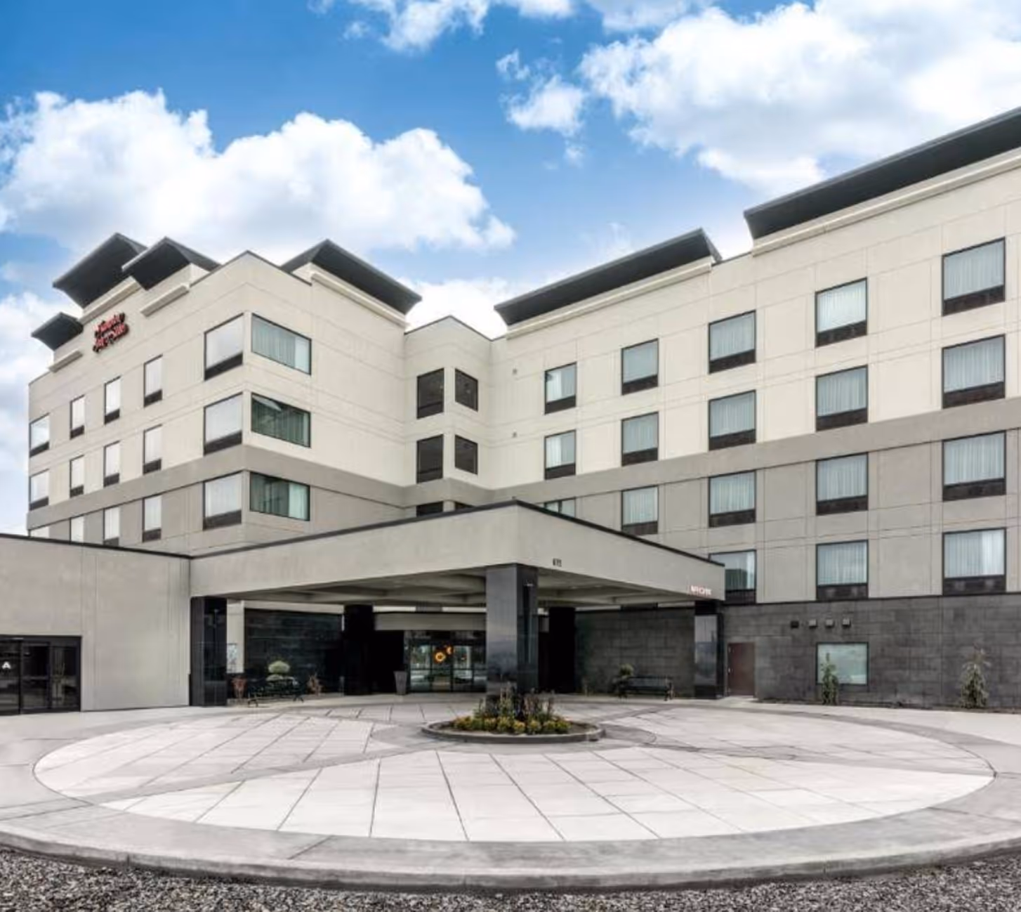 Modern multi-story hotel building with covered entrance and circular driveway under blue sky with clouds.