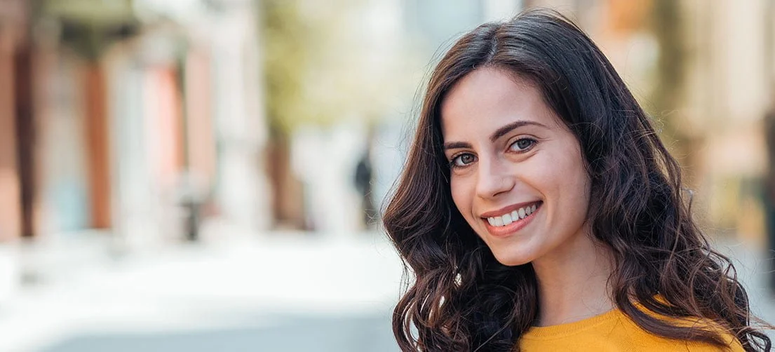 Smiling young woman with long dark hair wearing a yellow top outdoors in a blurred city background.