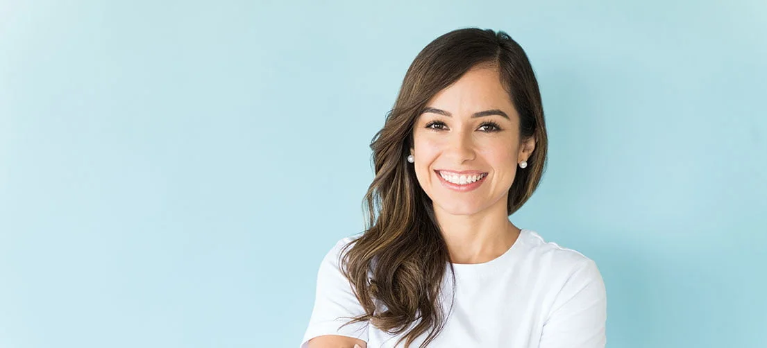 Smiling woman with long wavy brown hair wearing a white shirt against a light blue background.