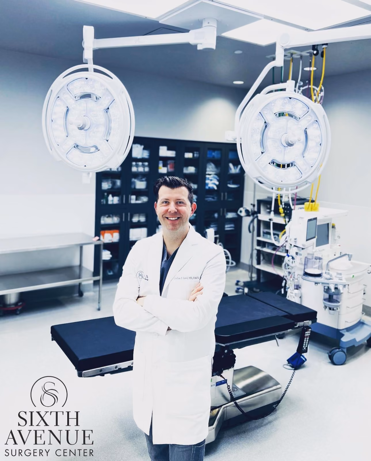 Smiling male doctor in white coat standing with arms crossed in a modern surgical operating room with overhead surgical lights and medical equipment.