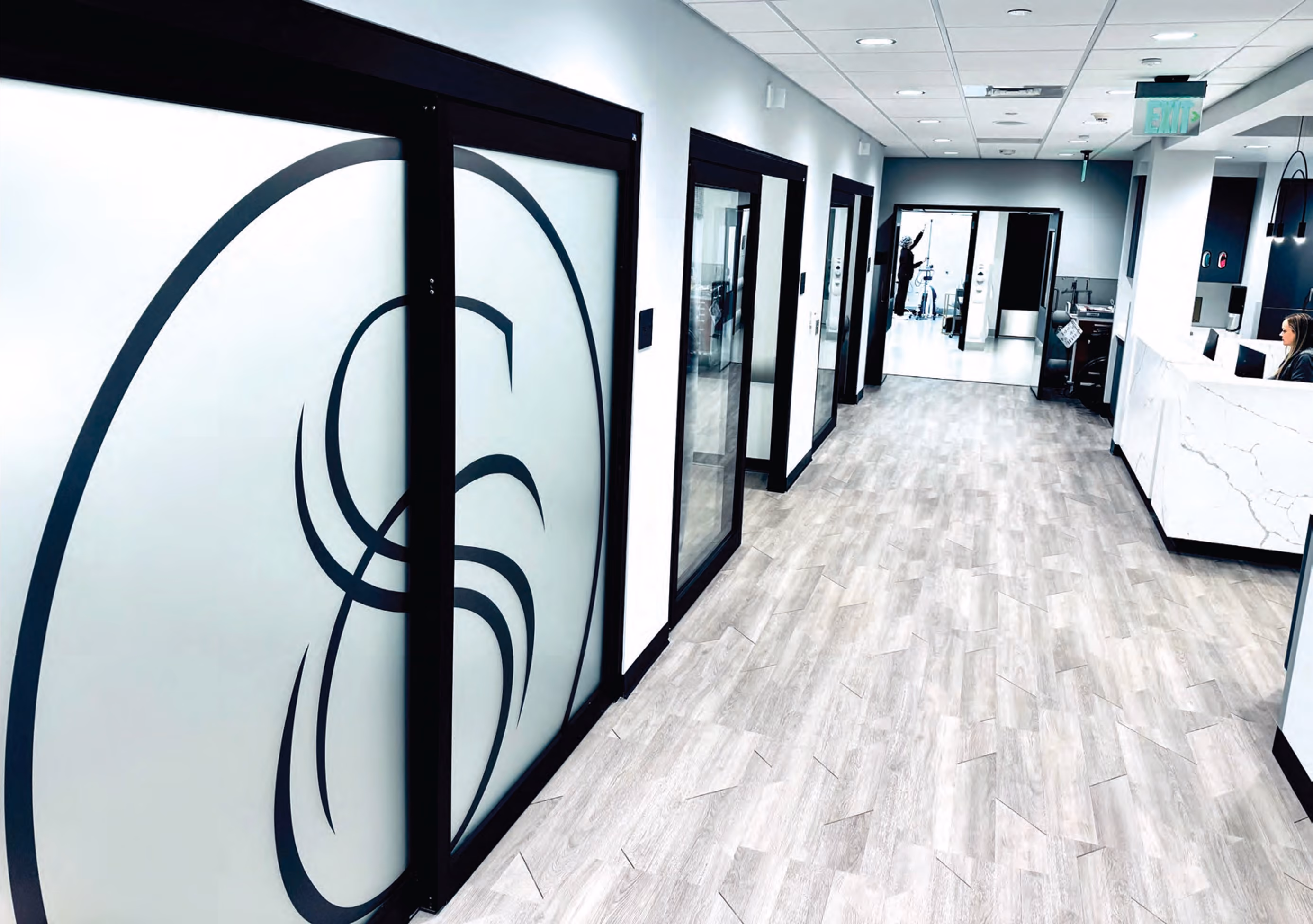 Modern office hallway with frosted glass doors featuring a circular abstract design, light wood flooring, and a white marble reception desk on the right with a seated receptionist.