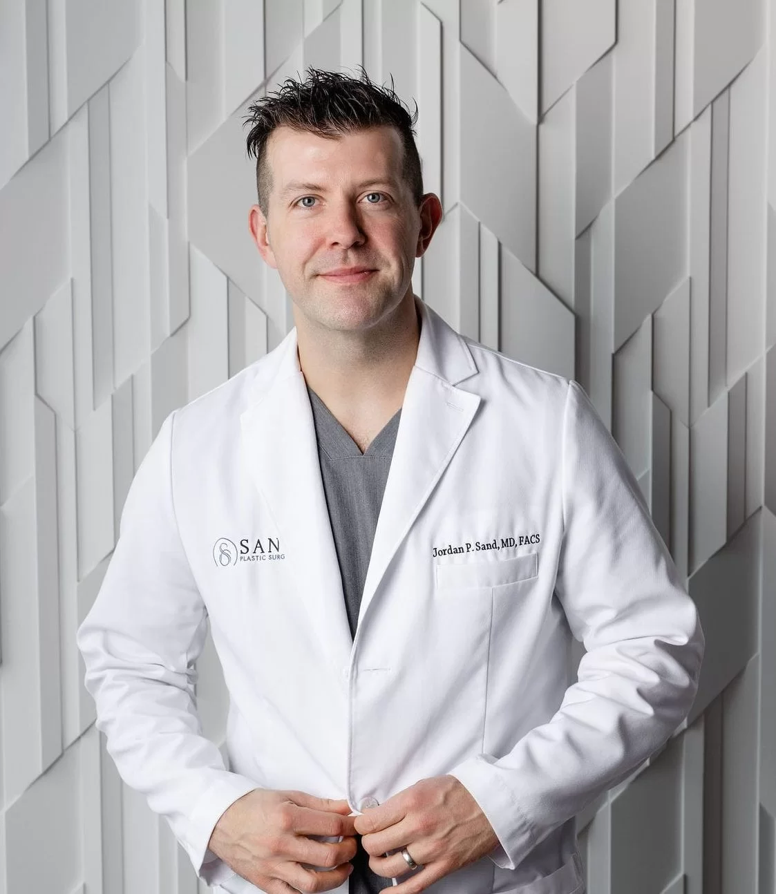Portrait of a man with short dark hair wearing a white coat over gray scrubs, standing against a gray geometric patterned wall, with the coat embroidered with SAN Plastic Surgery and Jordan P. Sand, MD, FACS.