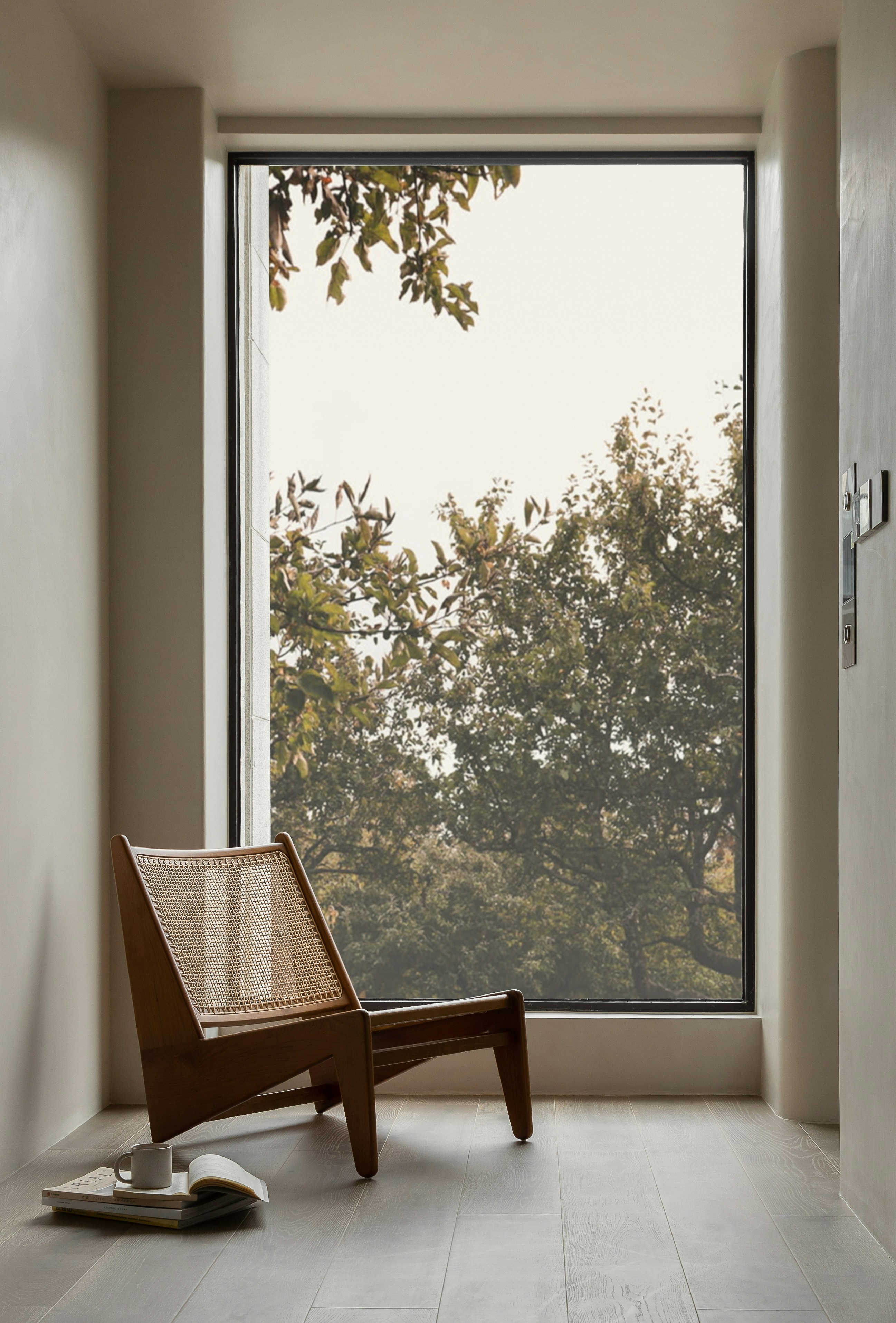 Wooden chair with woven back next to a large window overlooking green trees, with books and a cup on floor.